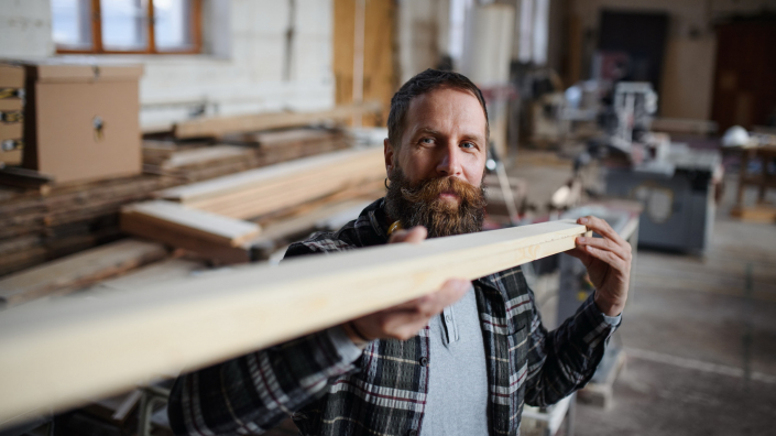Mature male carpenter carrying wooden board indoors in carpentery workshop. Small business concept.